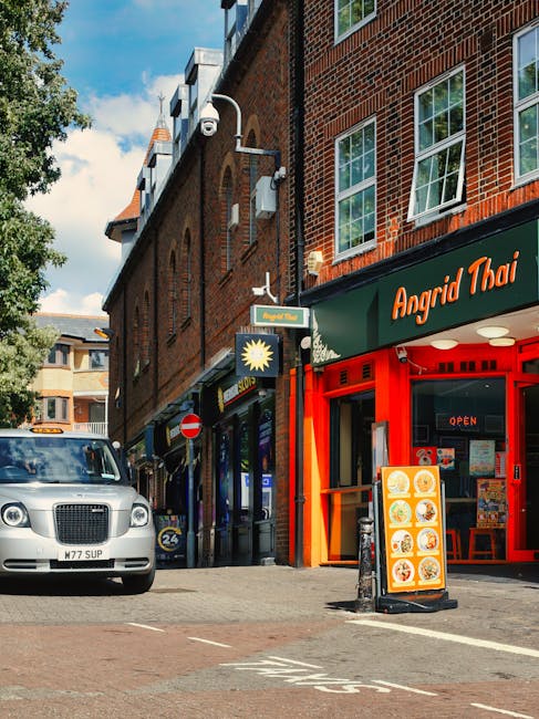 A street scene on Tooting High Street showing a busy urban environment with pedestrians crossing at a pedestrian traffic light, some carrying bags. Several vehicles, including cars and a van, are pres